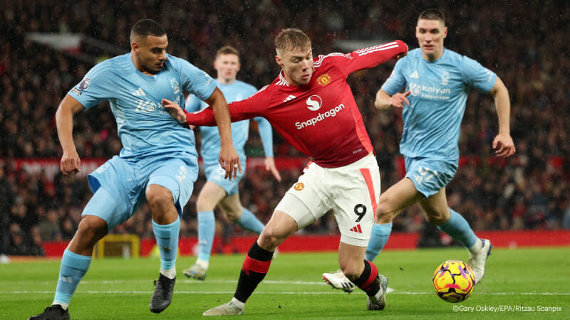 En fotbollsspelare från Manchester United, iklädd en röd matchtröja, kämpar om bollen med spelare från Nottingham Forest, klädda i ljusblått. Scenen utspelar sig på en regnig kväll under en Premier League-match, med intensiva rörelser och publik i bakgrunden.